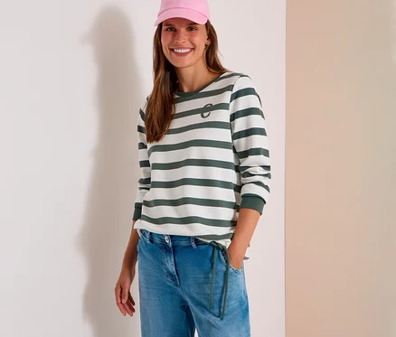 Lächelnde Frau mit rosa Kappe, einem weiß-grün gestreiften CECIL Sweatshirt mit „C“-Logo und hellblauen Jeans, die Hand in der Tasche.