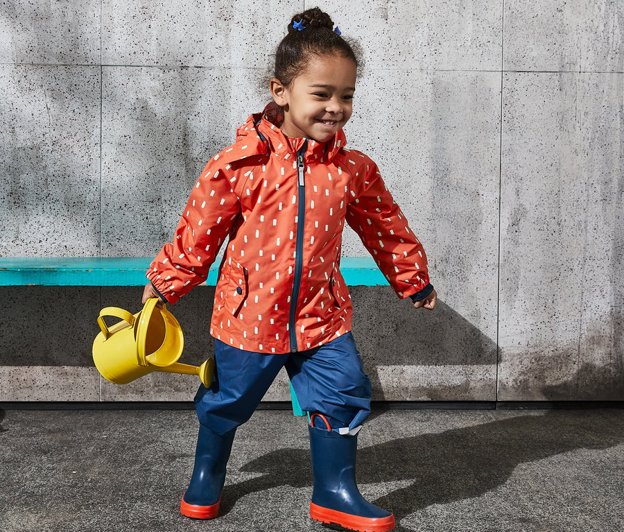 Mädchen trägt eine orange Regenjacke aus recyceltem Material mit Alloverprint und hält eine gelbe Gießkanne.