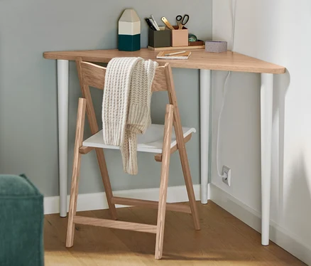 Ein Klappstuhl aus FSC®-zertifiziertem Eschenholz steht an einem dreieckigen Tisch. An der Wand hängt eine weiße Wandleuchte aus Metall.