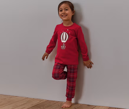 Lächelndes Mädchen steht an einer Wand, trägt einen roten Pyjama mit karierter Hose und einem Ballon-Aufdruck, der im Dunkeln leuchtet.
