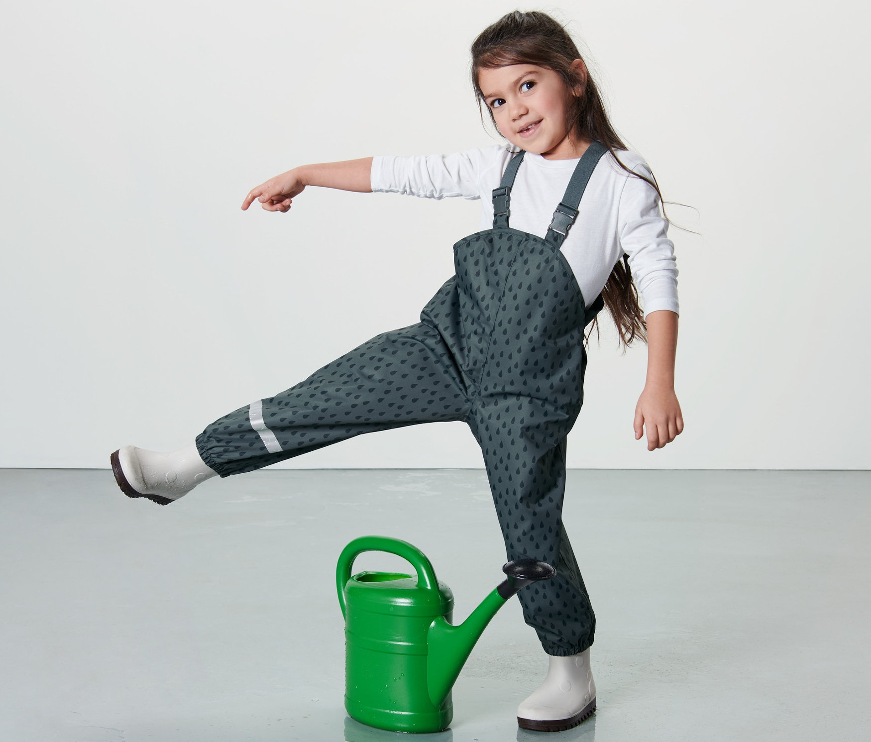 Mädchen steht mit einem Bein auf einer grünen Gießkanne und trägt eine Kleinkind-Regenhose, schieferblau.