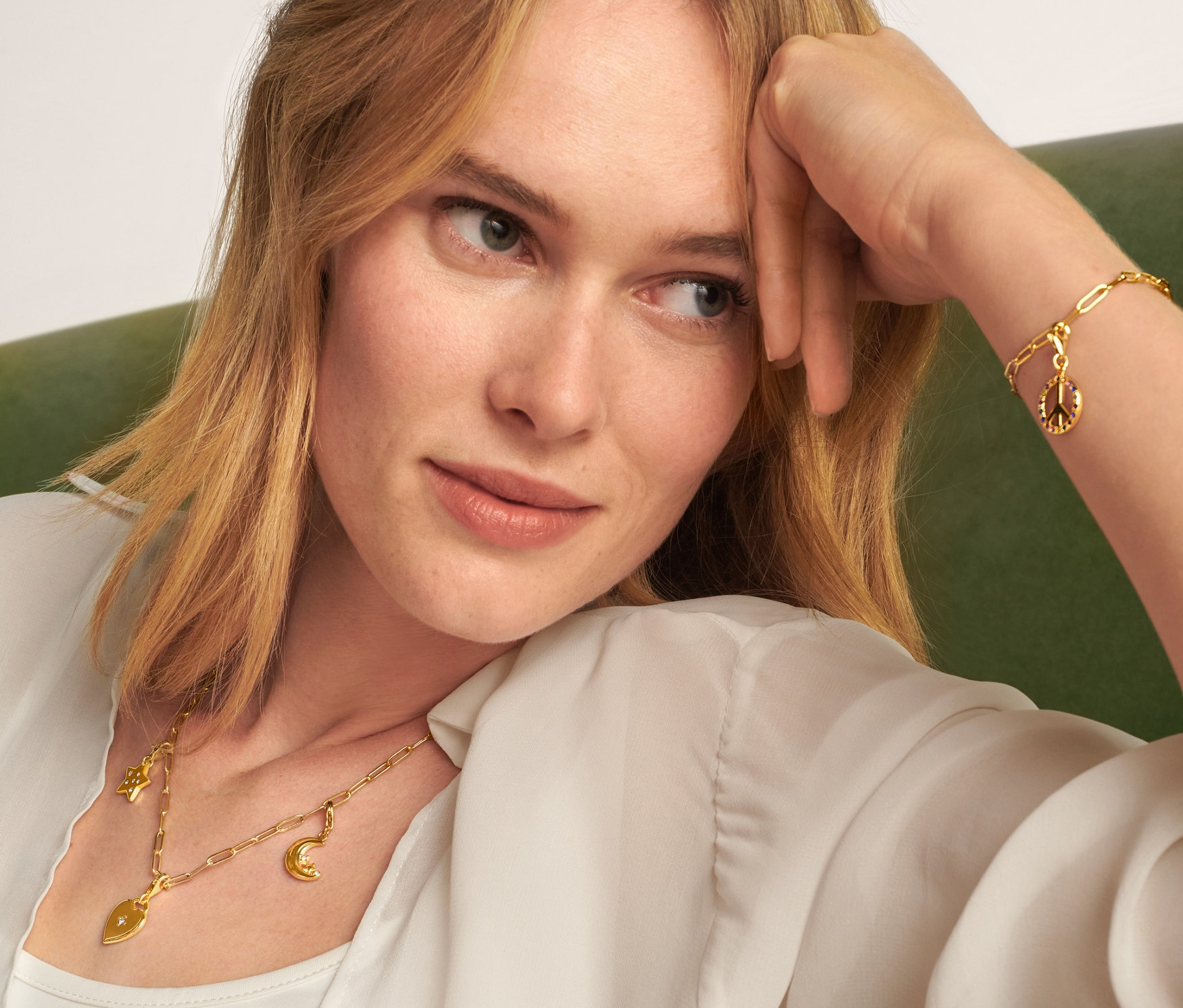 Blonde Frau sitzt auf einem grünen Sofa und trägt eine goldene Kette Collect, ein Armband Collect, ein Charm Set Love and Peace und ein Charm Set Moon and Stars.