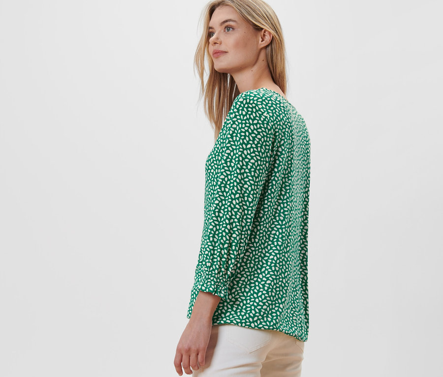 Blonde Frau posiert seitlich in einem grünen bedruckten Blusenshirt.
