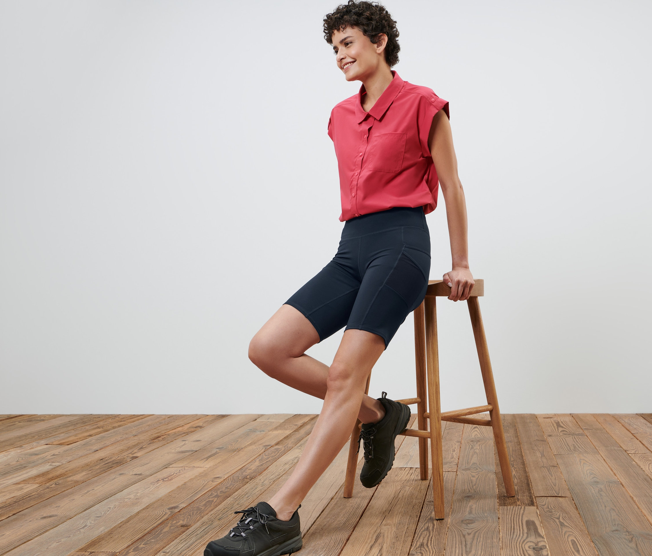 Frau sitzt auf einem Hocker und trägt Radlerhose, ein rotes Hemd und schwarze Schuhe.