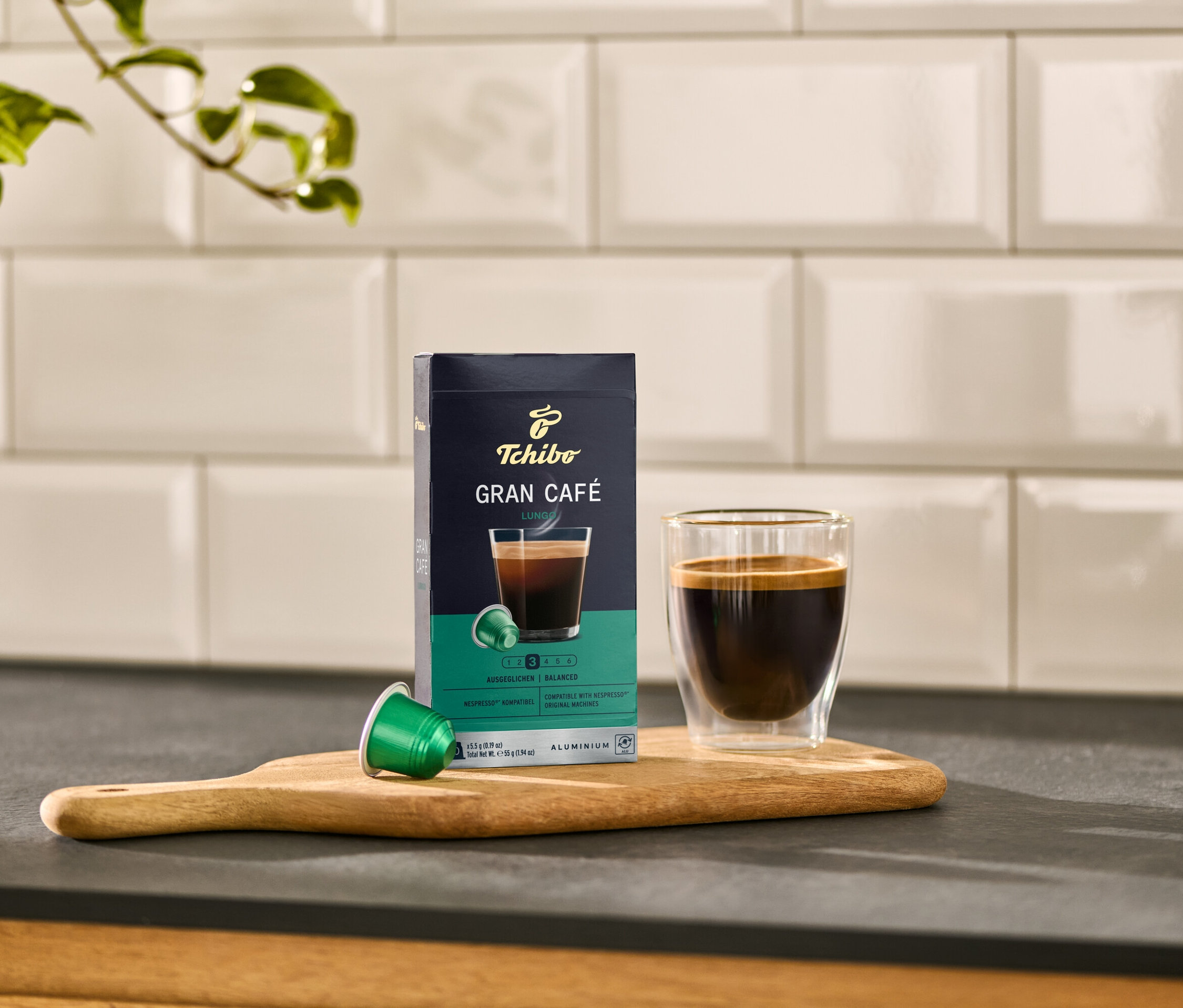 Auf einem Holzbrett stehen eine Packung Gran Café Lungo, eine grüne Kapsel und ein Glas Kaffee.