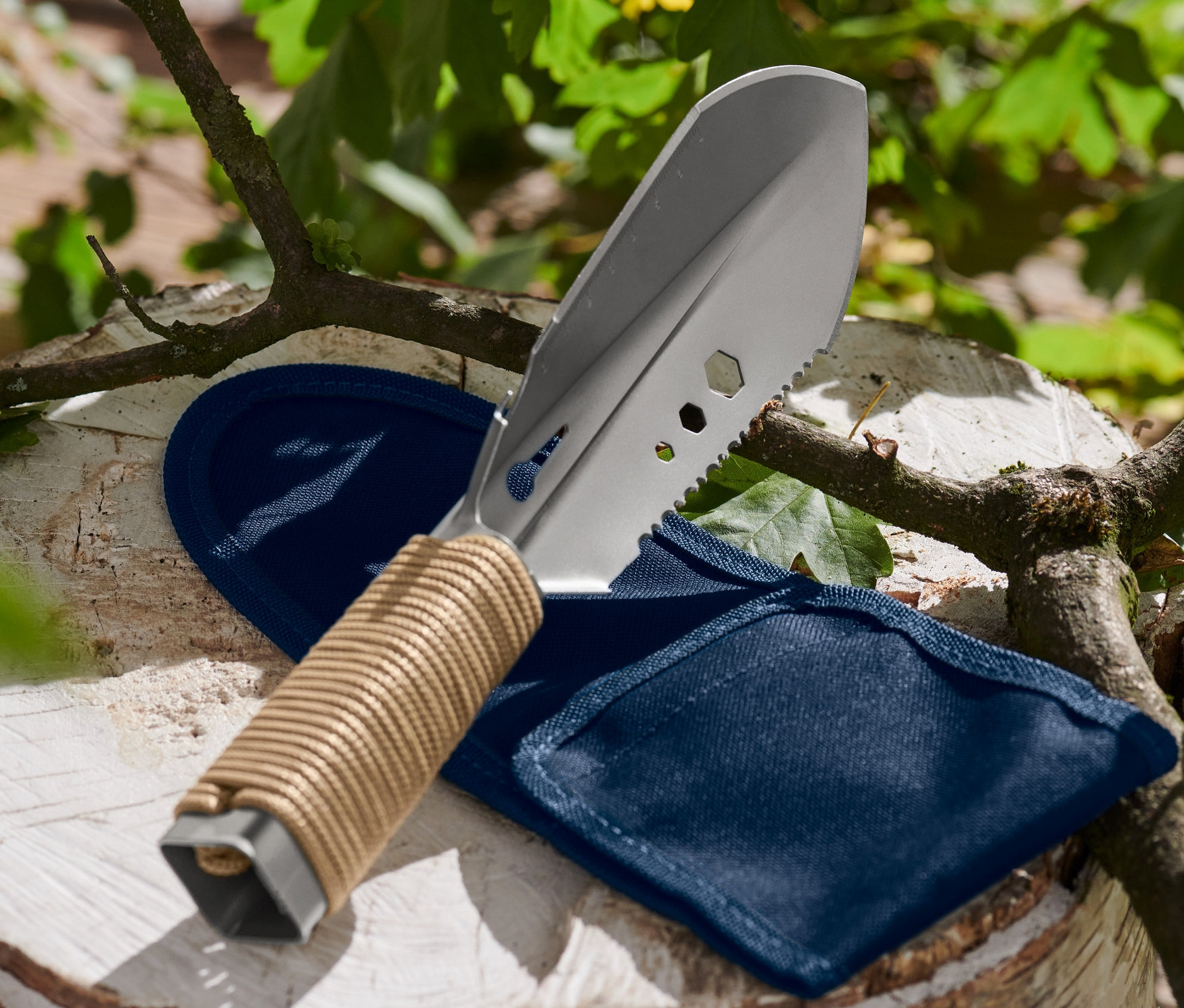 Eine 9-in-1-Camping-Schaufel liegt neben einer blauen Hülle auf einem Holzscheit.