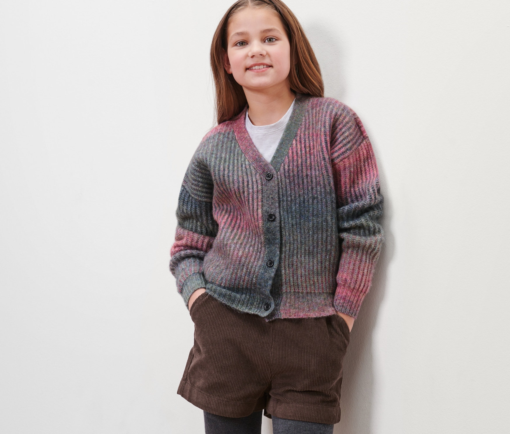 Mädchen trägt eine Kinder-Strickjacke mit Schurwolle, braune Shorts und graue Leggings.