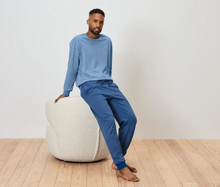 Mann sitzt auf einem weißen Pouf und trägt ein blaues Langarmshirt und eine blaue Jogginghose.
