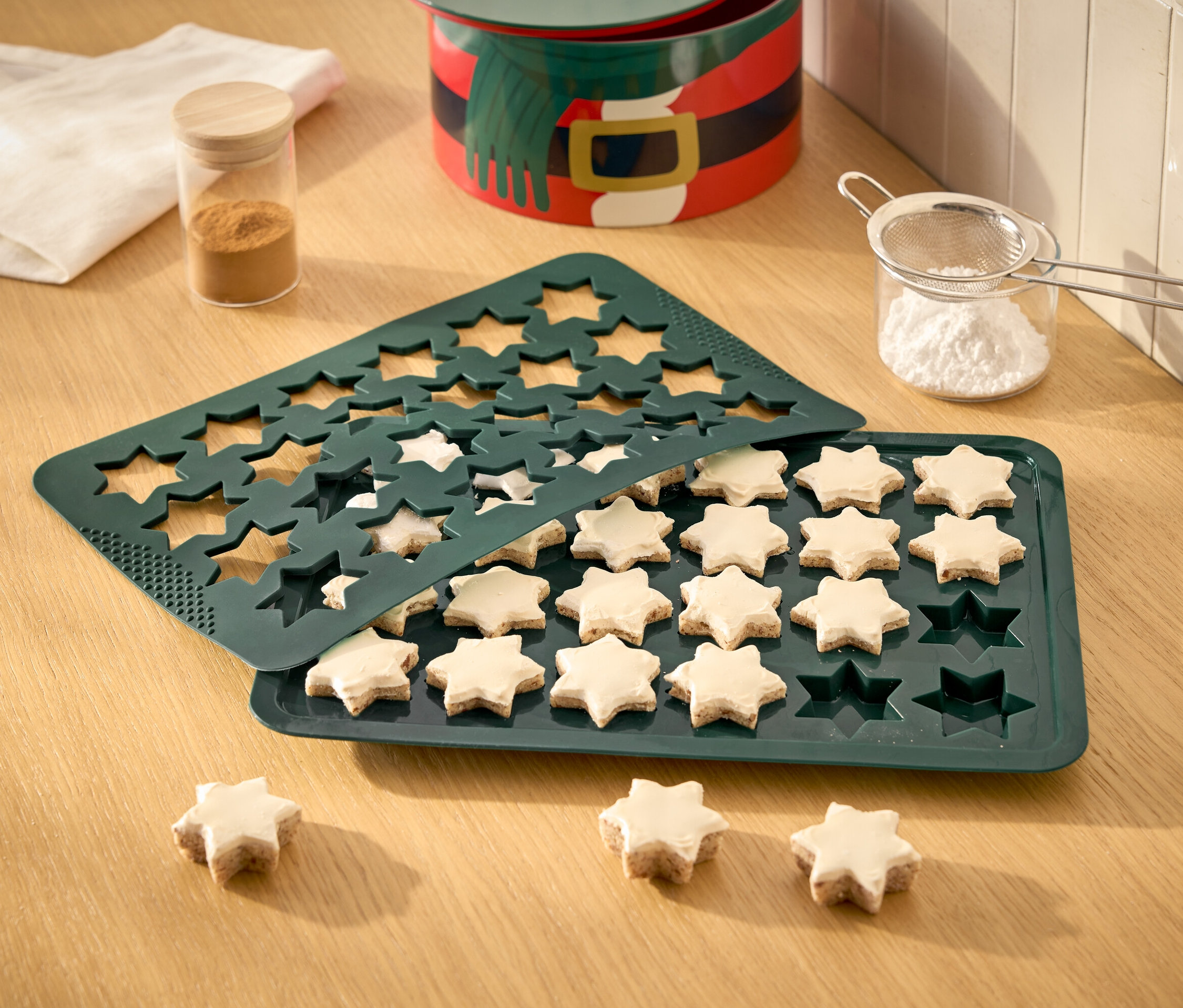 Sterne in einer grünen Backform, einige sind bereits gebacken und mit Zuckerguss verziert, Puderzuckersieb, Dose und Geschirrtuch auf einem Holztisch.