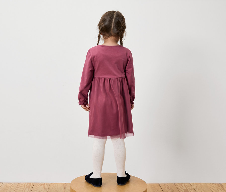 Mädchen in einem burgunderroten Langarmkleid, weiß gemusterten Strumpfhosen und schwarzen Schuhen steht mit dem Rücken zur Kamera.