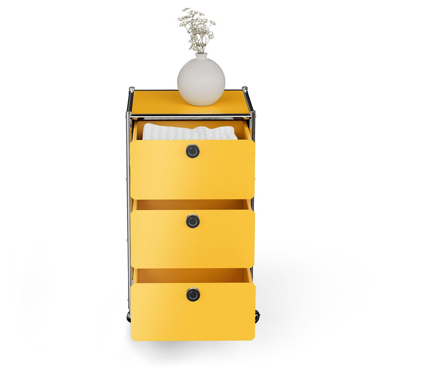 Gelbe Kommode mit drei Schubladen, die oberste Schublade ist offen und enthält weiße Kleidung, auf der Kommode steht eine weiße Vase mit weißen Blumen.