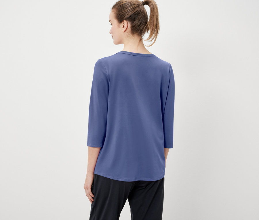 Rückansicht einer Frau mit zum Pferdeschwanz hochgebundenen Haaren, die ein blaues Dreiviertelarm-Shirt und eine schwarze Hose trägt.
