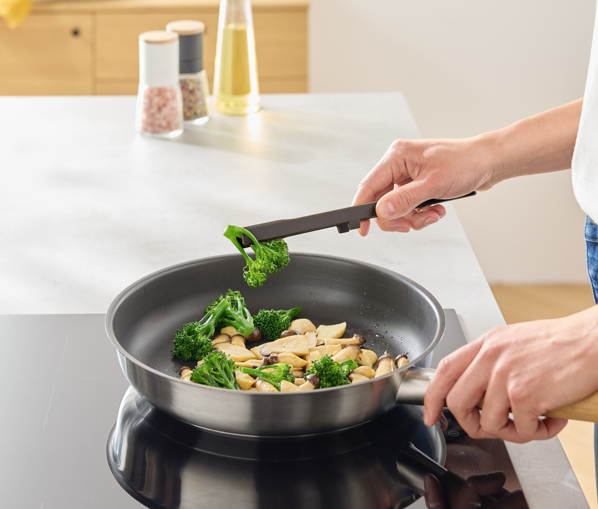 Person fügt mit einer Zange Brokkoli in eine Pfanne mit Pilzen.