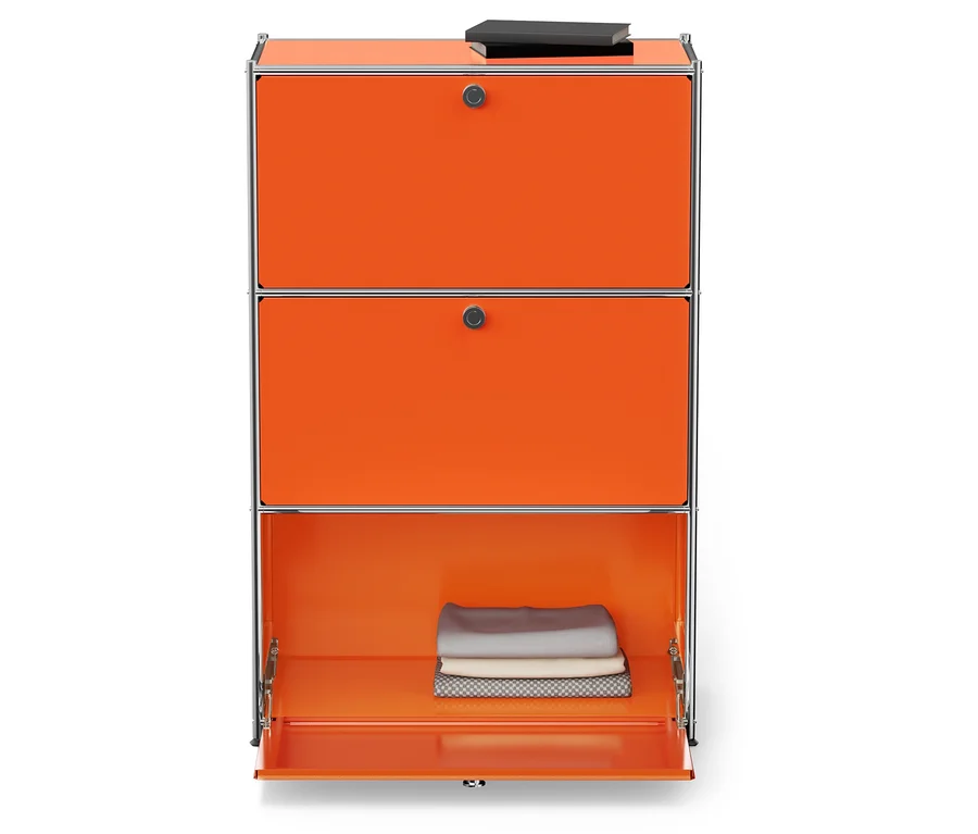 Orangefarbener Schrank mit silbernem Rahmen, die untere Tür ist geöffnet und innen liegen gefaltete Stoffe. Oben liegt ein Buch.