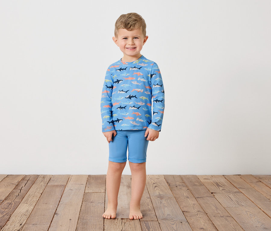 Ein kleiner Junge steht barfuß auf einem Holzboden und trägt ein blaues Langarmshirt mit Hai-Muster und blaue Shorts.
