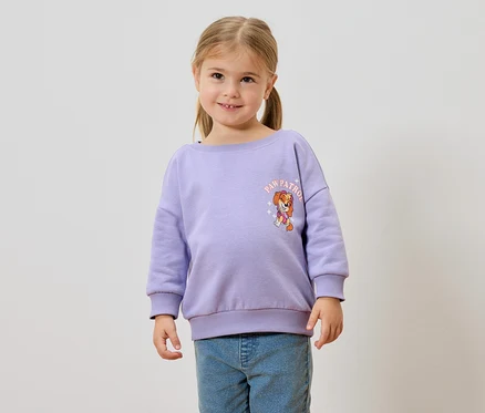 Mädchen mit Zöpfen trägt einen lila Pullover mit Paw Patrol Aufdruck und blaue Jeans.