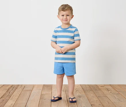 Ein Junge mit blonden Haaren steht barfuß auf einem Holzboden und trägt ein gestreiftes blau-weißes T-Shirt, blaue Shorts und blaue Flip-Flops.