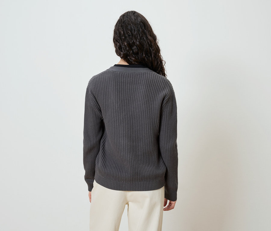 Rückansicht einer Frau mit langen, lockigen, dunklen Haaren, die einen grauen Strickpullover und eine cremefarbene Hose trägt.