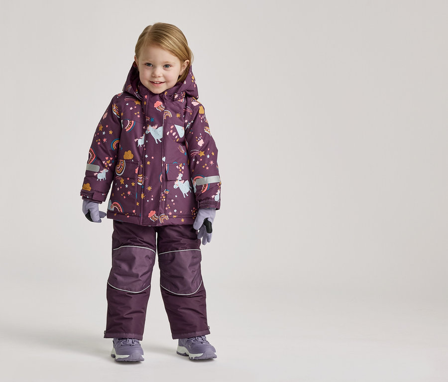 Ein kleines Mädchen steht in einem lila Schneeanzug mit Einhorn- und Regenbogenmuster und grauen Handschuhen und Schuhen vor einem hellen Hintergrund.