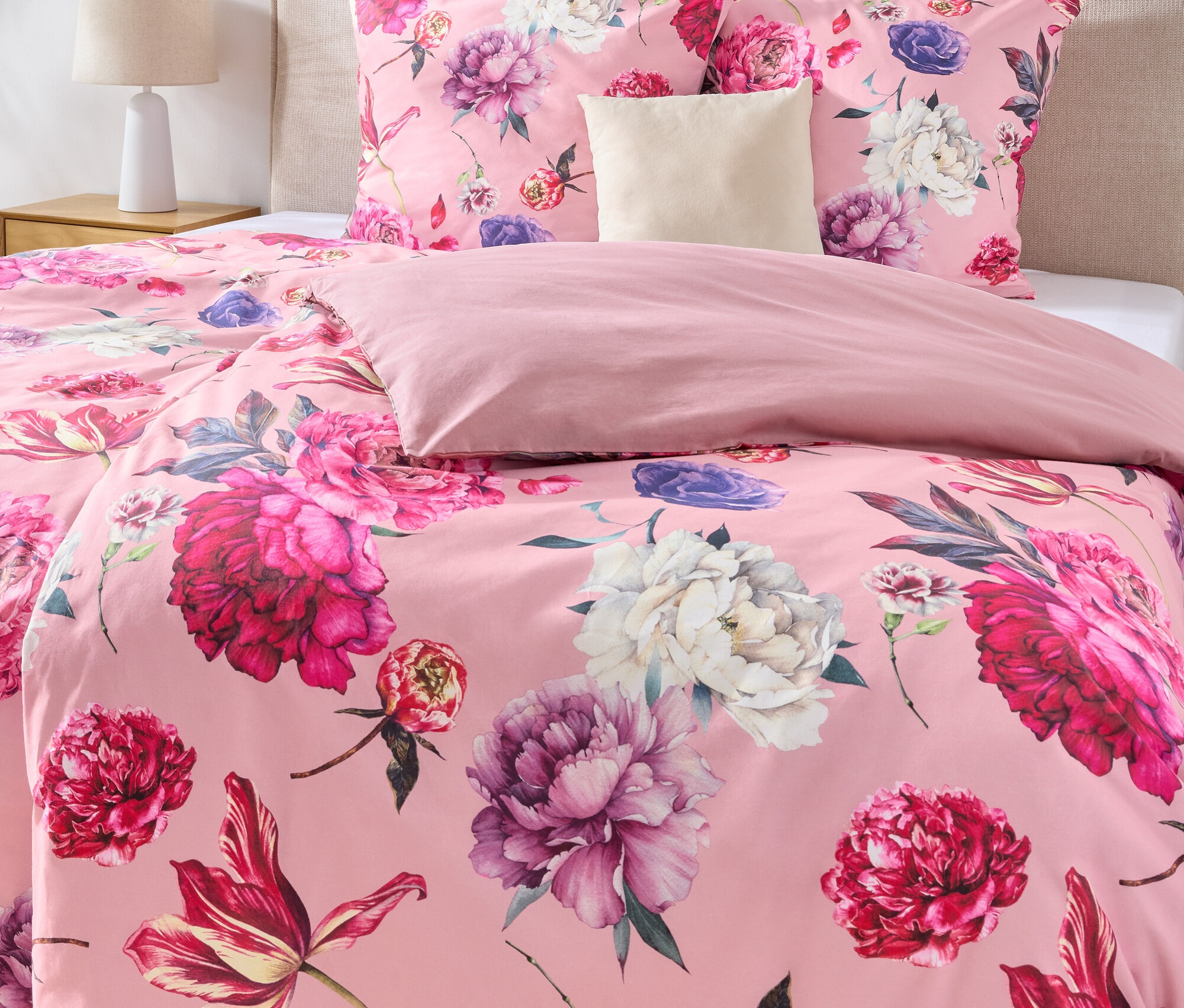 Nahaufnahme eines Bettes mit rosa Bettwäsche mit Blumenmuster, einem beigen Kopfteil und zwei gerahmten Bildern an der Wand.