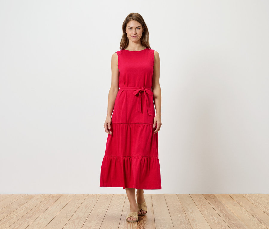 Eine Frau steht vor einer weißen Wand und trägt ein rotes, ärmelloses, gestuftes Kleid mit Gürtel und Sandalen.