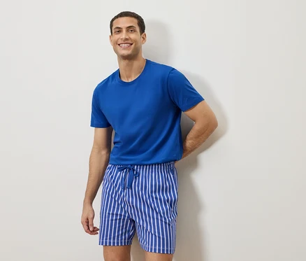 Mann mit blauem T-Shirt und gestreiften Shorts steht vor einer weißen Wand.