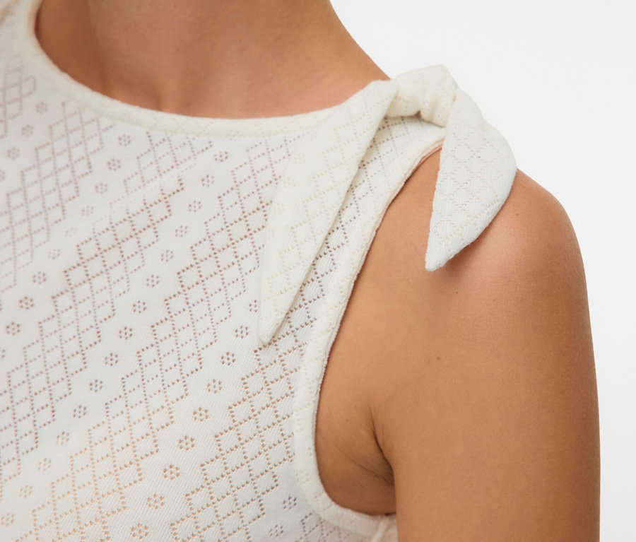 Nahaufnahme einer Frau, die ein ärmelloses, weißes Lochstickereioberteil mit einer Schleife an der Schulter trägt.