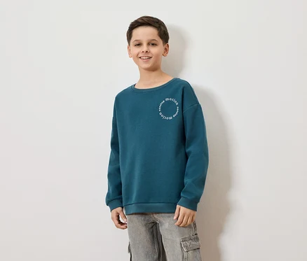 Ein lächelnder Junge trägt ein blaugrünes Sweatshirt mit der Aufschrift "nonstop motion" und graue Cargohosen vor einem weißen Hintergrund.
