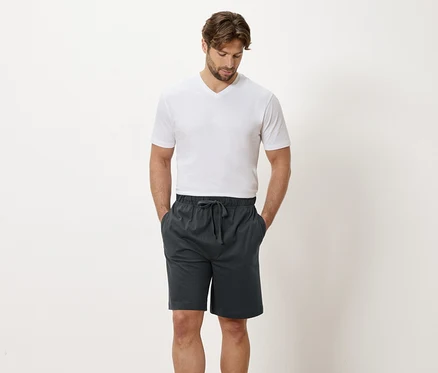 Mann mit weißem T-Shirt und grauen Shorts steht vor einer weißen Wand.