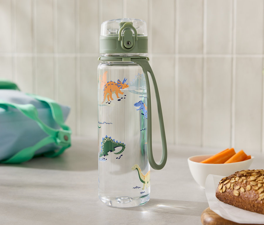 Eine durchsichtige Wasserflasche mit Dinosaurier-Motiven steht neben einem Brot und einer Schüssel Karotten.