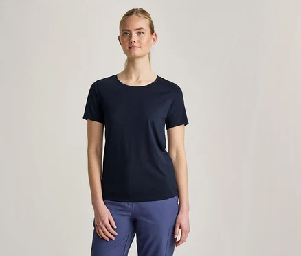 Eine Frau mit zurückgebundenem blondem Haar trägt ein dunkelblaues Kurzarm-T-Shirt und blaue Hosen, während sie vor einem hellgrauen Hintergrund steht.