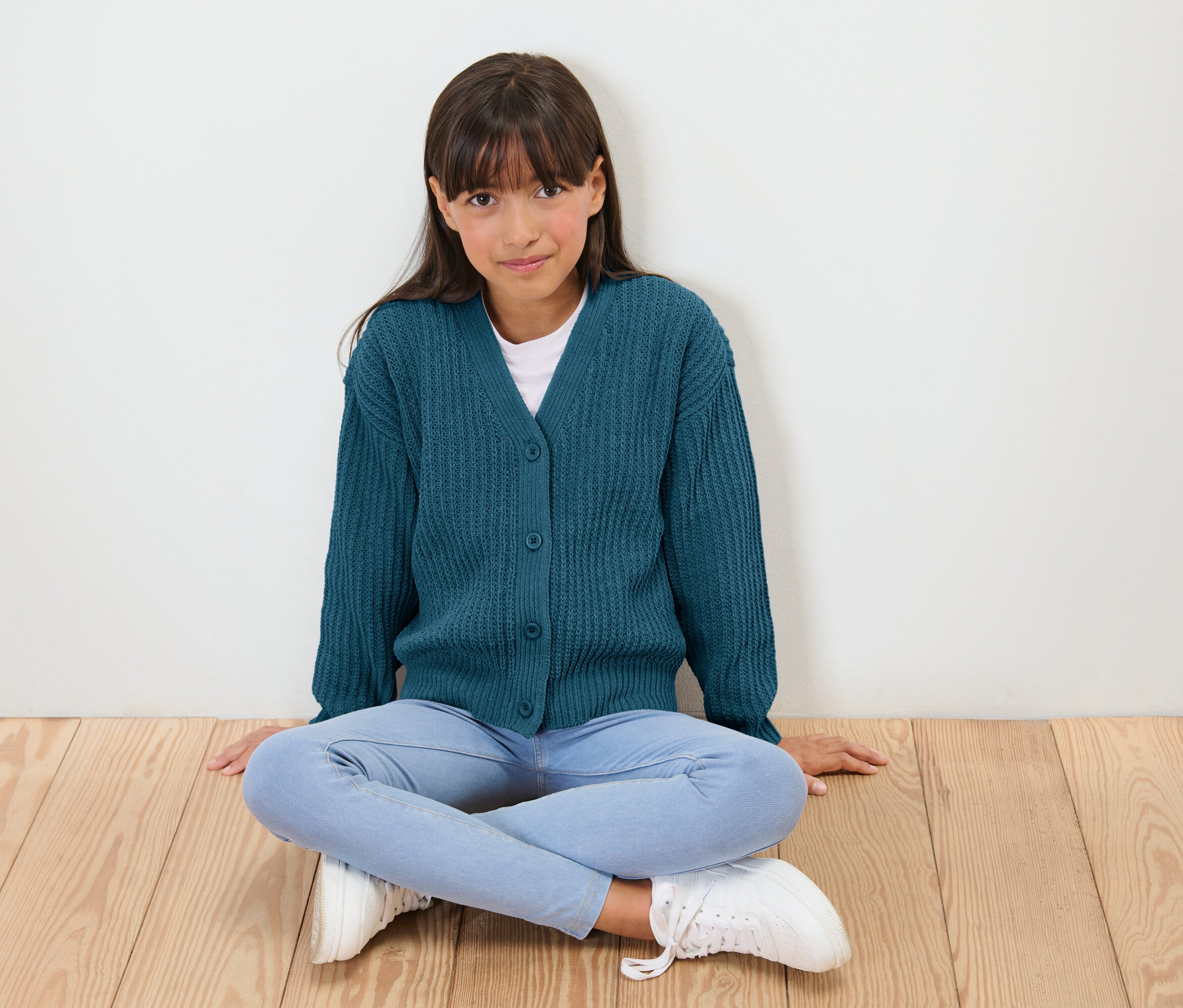 Ein Mädchen sitzt mit gekreuzten Beinen auf einem Holzboden, gekleidet in eine blaue Strickjacke, eine weiße Bluse, Jeans und weiße Turnschuhe.