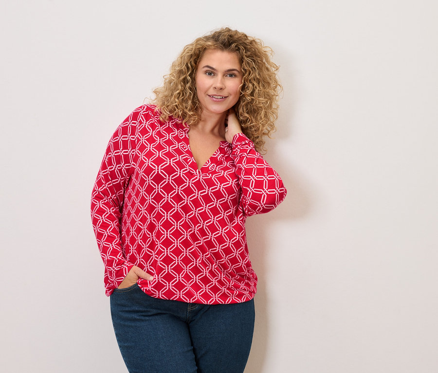 Frau mit lockigen Haaren, die eine rot-weiße Bluse und Jeans trägt und mit einer Hand in der Hosentasche posiert.