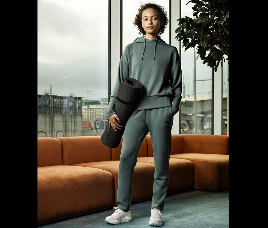 Junge Frau in grauer Trainingsbekleidung hält eine zusammengerollte Yogamatte vor einem Fenster.