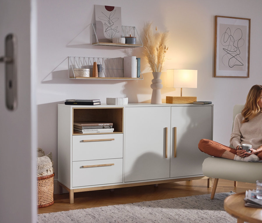 Frau sitzt im Wohnzimmer neben einem weißen Sideboard »Eklund« mit Schubladen und Tür.