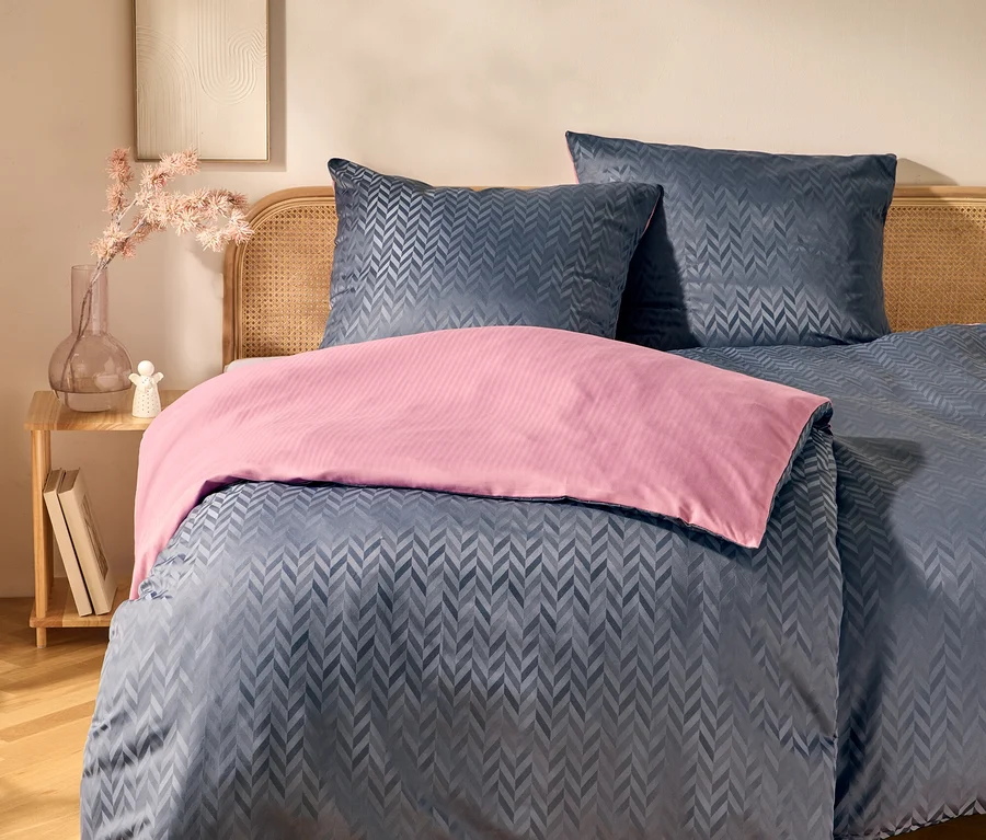 Bett mit Rattan-Kopfteil, dunkelblauer Bettwäsche mit Chevron-Muster und rosa Überwurf. Daneben ein Holztisch mit Vase, rosa Blumen und Büchern. An der Wand hängt ein gerahmtes Kunstwerk.