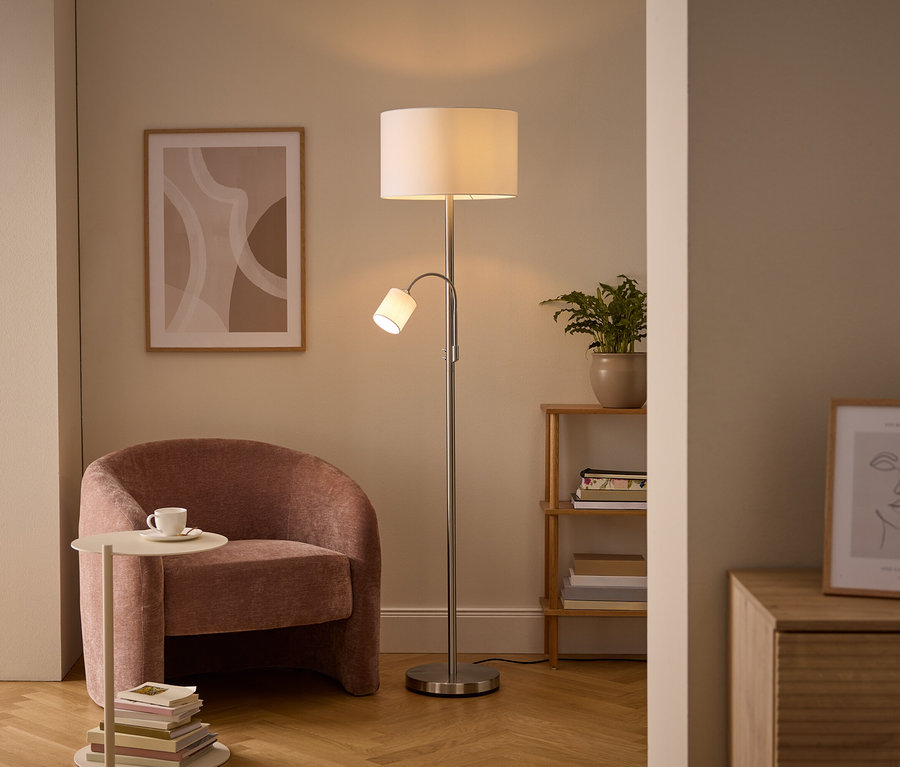 Gemütliche Wohnzimmerecke mit einer silbernen Stehlampe mit Haupt- und Leselicht, einem rosa Polstersessel und einem Beistelltisch mit Tasse. Wandkunst und ein Holzregal mit Pflanze sind ebenfalls sichtbar.