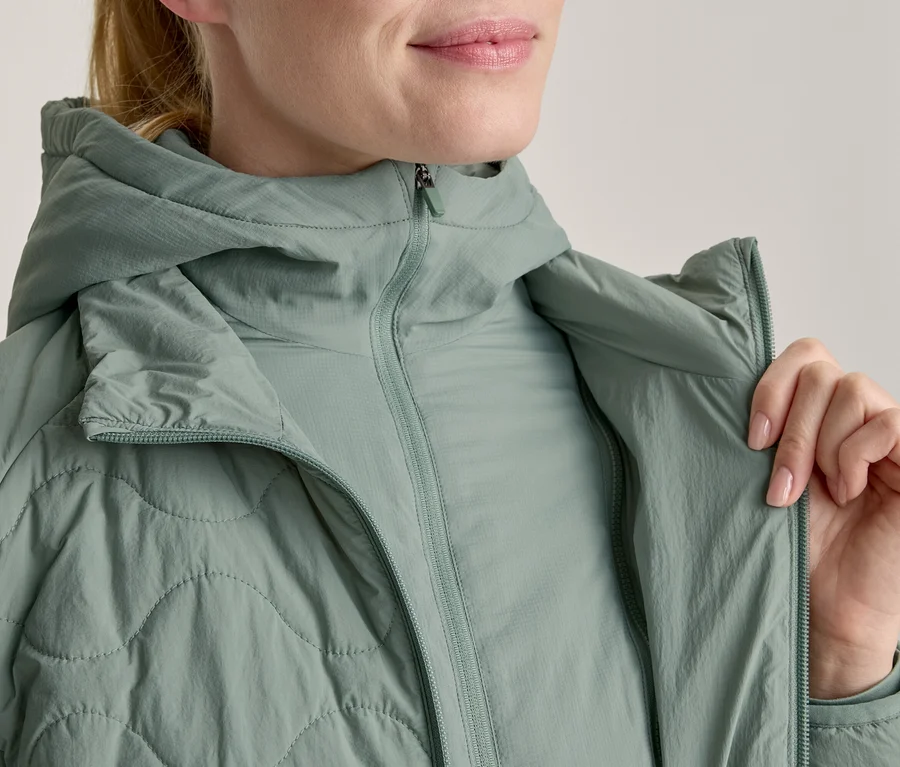 Nahaufnahme einer Frau, die eine hellgrüne, gesteppte Kapuzenjacke über einem helleren grünen Langarmshirt trägt. Ihre Hand hält das Revers der Jacke, ihr Mund lächelt leicht.