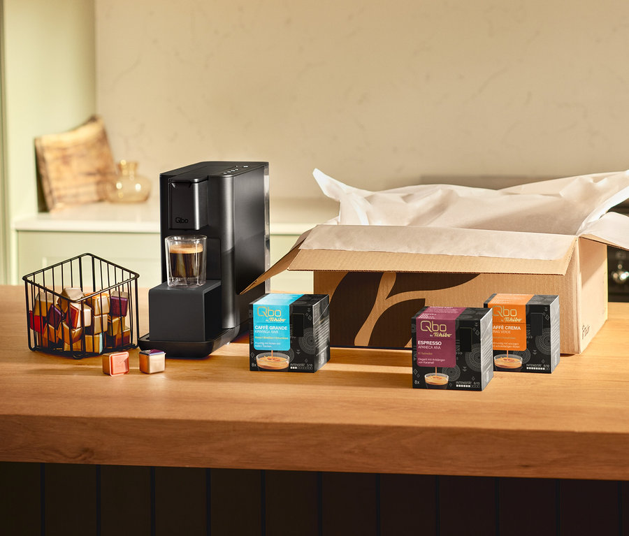 Eine Qbo Iconic, Dark Chrome Kaffeemaschine mit einem Glas Kaffee darunter steht auf einer Holztheke. Daneben ein Korb mit Qbo Kapseln und zwei einzelnen Kapseln. Rechts daneben stehen eine offene Qbo Caffè Grande KIRINYAGA BAYA Kapselpackung, eine Qbo Espresso APANECA ANA Kapselpackung und eine weitere Qbo Kapselpackung.