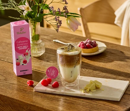 Auf einem Holztisch stehen ein Glas Kaffee, weiße Schokoladenstücke und Himbeeren auf einem Marmorbrett. Im Hintergrund ist eine Packung Cafissimo Flavoured Espresso - White Choc & Raspberry - 10 Kapseln zu sehen.