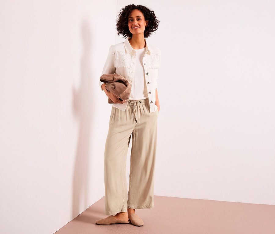 Lächelnde Frau steht, trägt ein weißes T-Shirt, eine weiße Jacke und eine beige Street One 7/8-Culotte, hält eine braune Clutch.