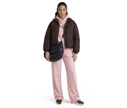 Eine Frau steht frontal, trägt eine dunkelbraune Pufferjacke, einen hellrosa Hoodie, hellrosa weite Jogginghosen und schwarz-leopard gemusterte Sneaker. Sie trägt eine schwarze gesteppte Umhängetasche.