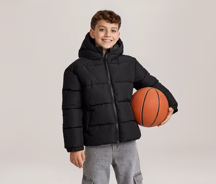 Ein lächelnder junger Junge in einer schwarzen Kapuzen-Steppjacke und grauen Jeans hält einen orangefarbenen Basketball unter seinem linken Arm.