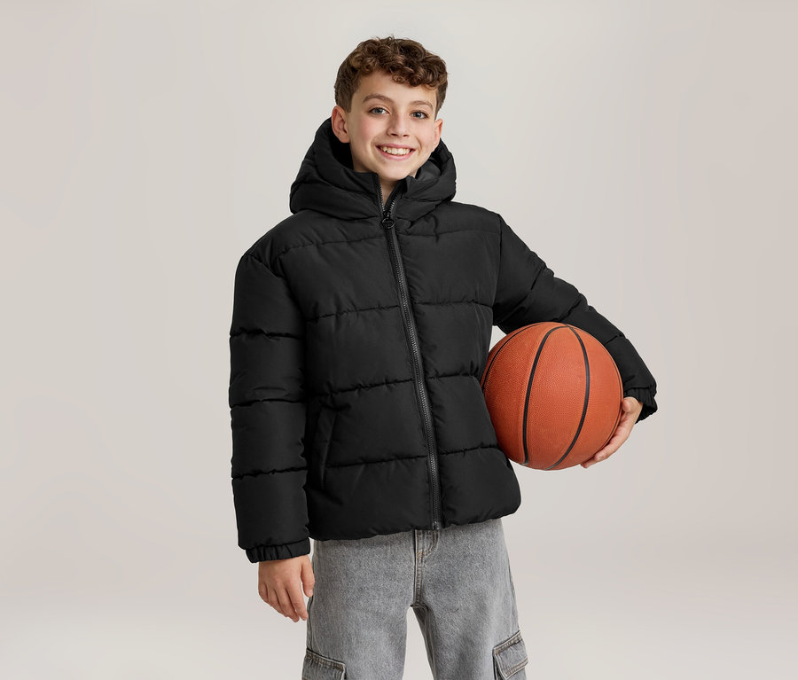 Ein lächelnder junger Junge in einer schwarzen Kapuzen-Steppjacke und grauen Jeans hält einen orangefarbenen Basketball unter seinem linken Arm.