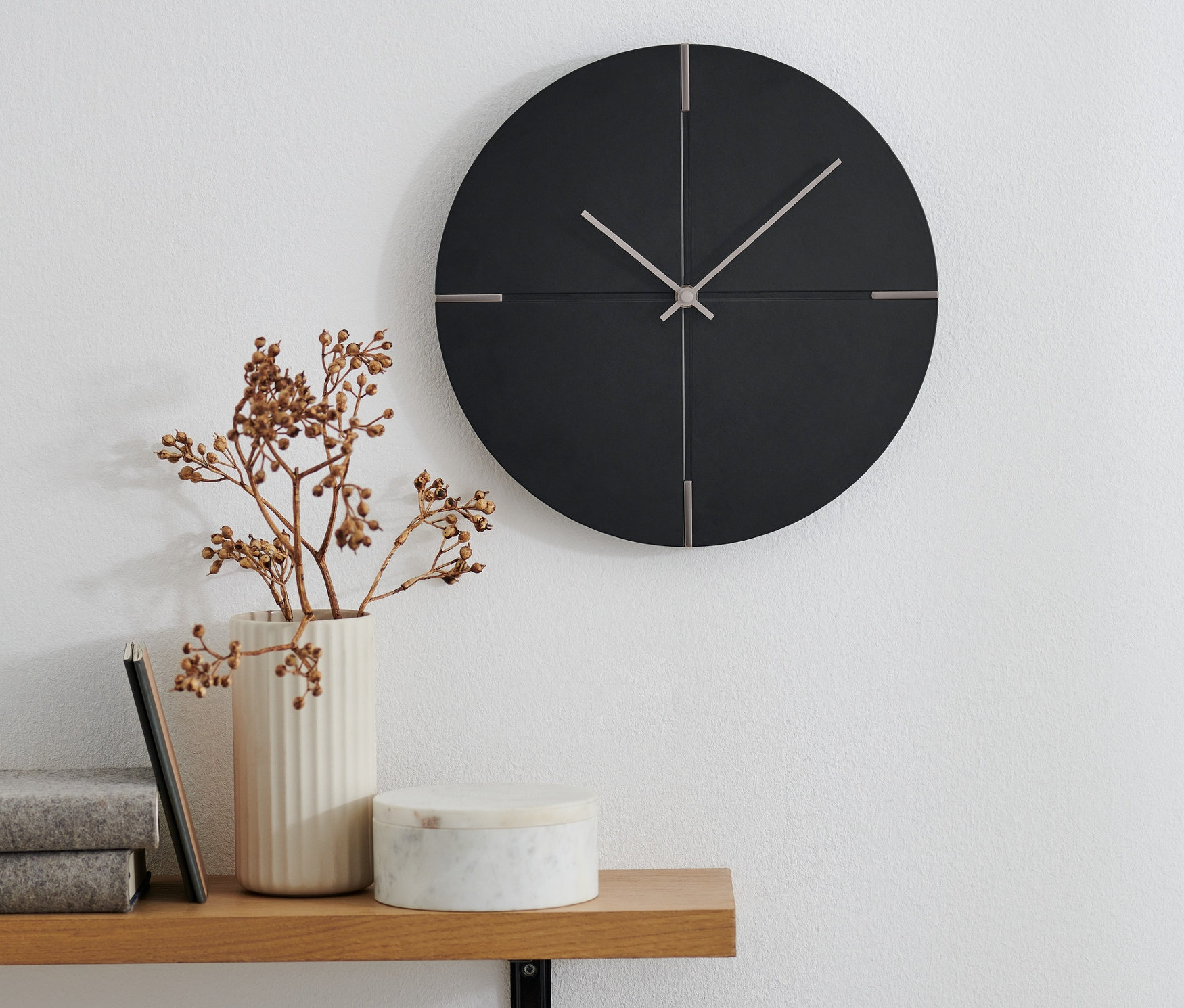 Eine Design-Wanduhr hängt an der Wand. Auf einem Regal darunter steht eine Vase mit Zweigen.