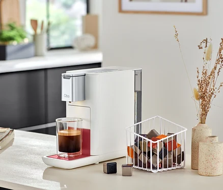 Qbo ICONIC Dark Chrome Kaffeemaschine neben Qbo ESSENTIAL Bright Coral mit Tasse Kaffee und Kapseln.