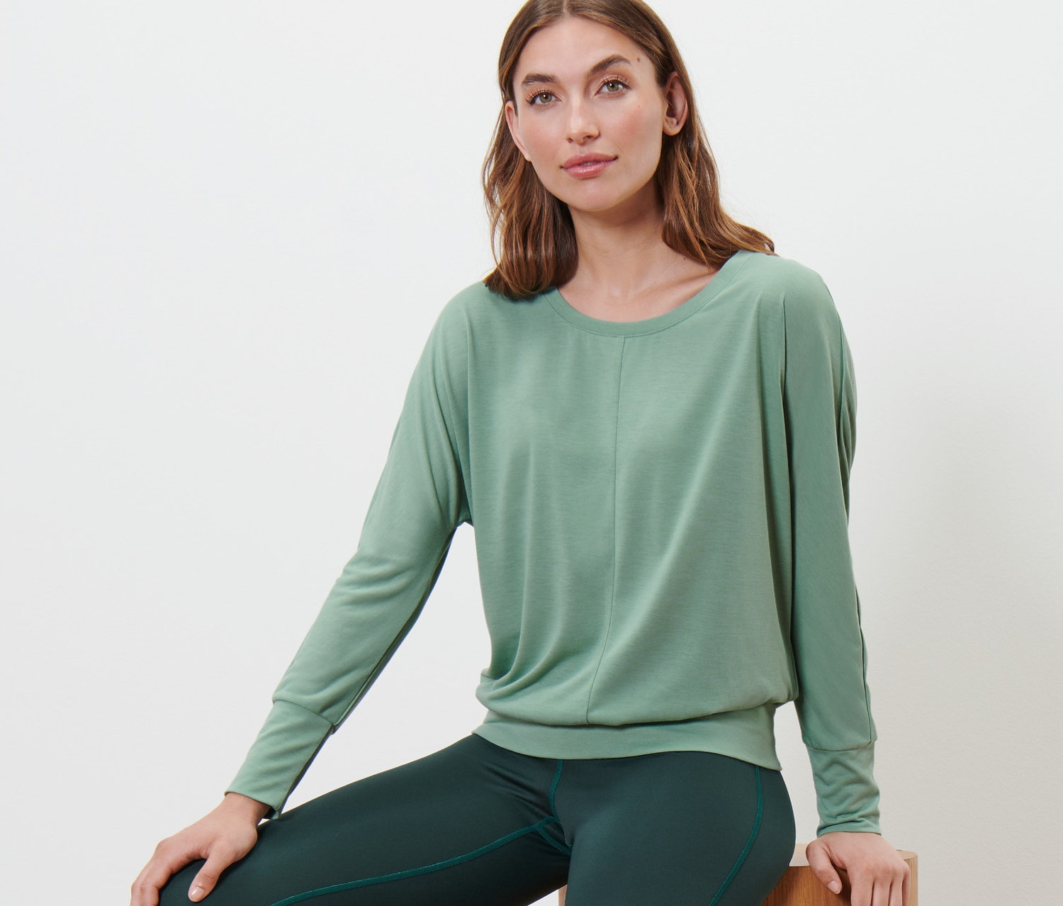 Frau sitzt auf Holzhockern und trägt ein Sport-und-Yogashirt in Salbeigrün.
