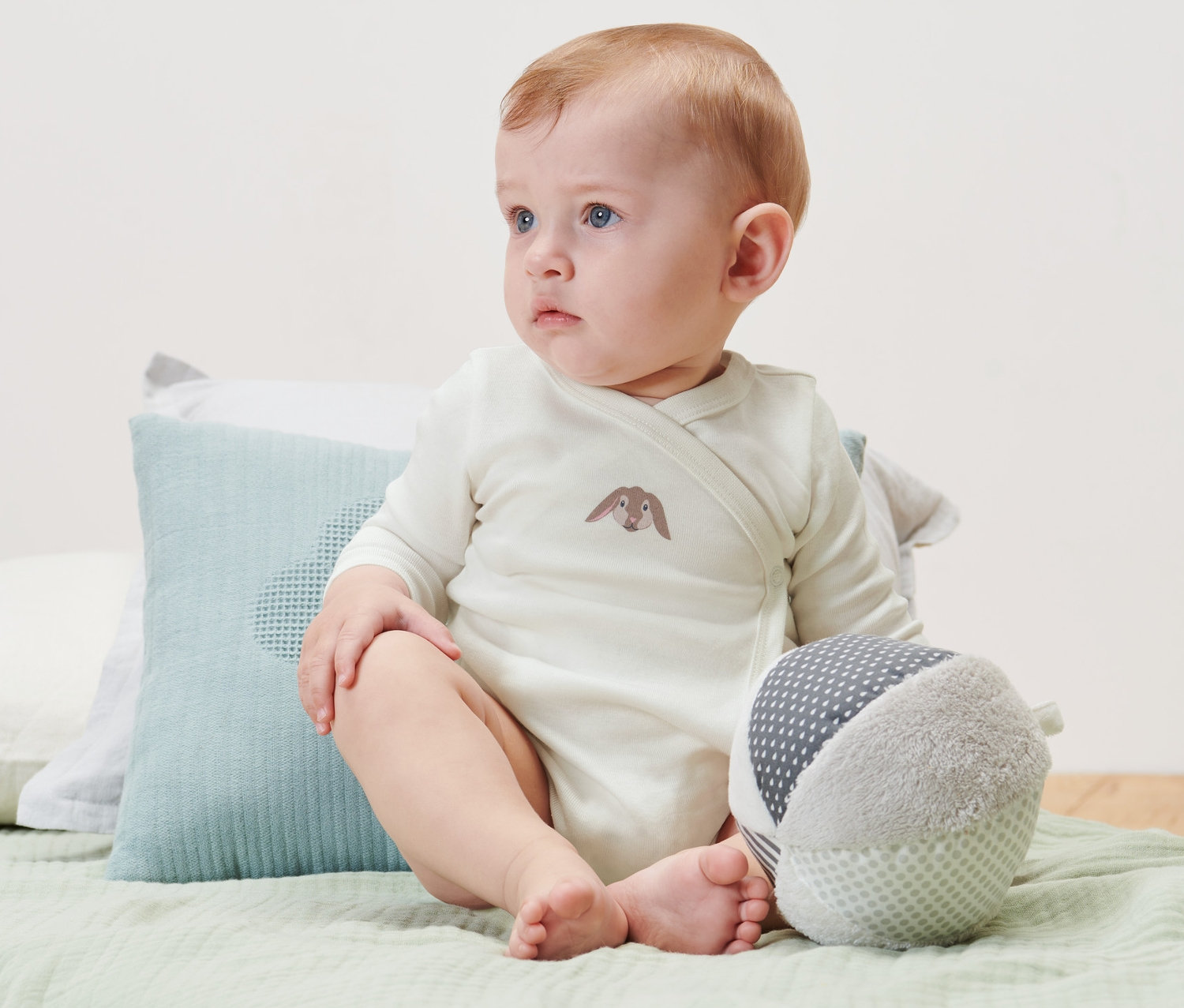 Baby sitzt auf einem Bett und trägt 3 Baby-Langarmbodys.