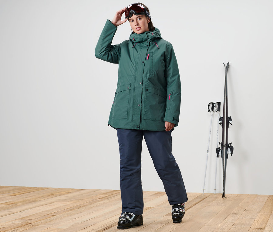 Frau posiert in Ski-und-Snowboardjacke und Skihose. Ski stehen neben ihr.