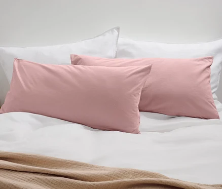 Zwei rosafarbene Jersey-Kopfkissenbezüge auf einem Bett mit weißen Kissen und einer beigen Decke.